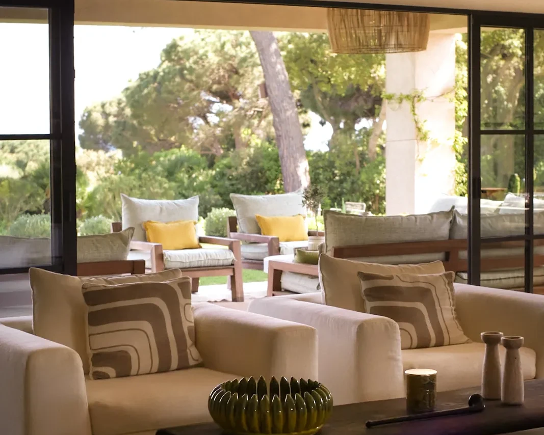 Un salon de villa ouvert sur la terrasse, portes repliées, pins parasols au fond